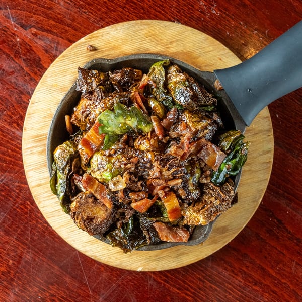 Crispy Brussels sprouts with bacon bits served in a cast iron skillet on a wooden board.