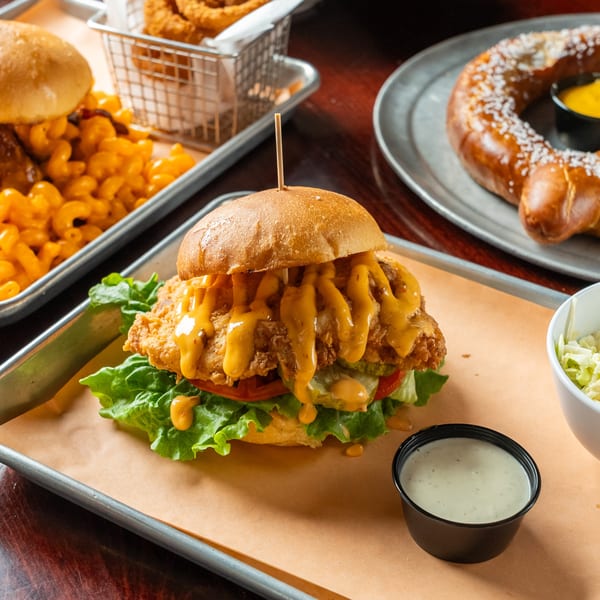 A fried chicken sandwich with cheese sauce, coleslaw, mac and cheese, and a pretzel with mustard.