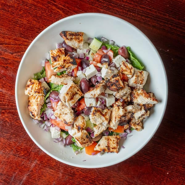 Overhead view of a grilled chicken salad with feta, olives, tomatoes, and red onion.