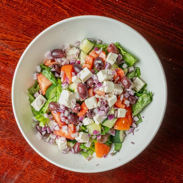 A vibrant Greek salad in a white bowl, featuring lettuce, tomatoes, cucumbers, olives, feta, and red onions.
