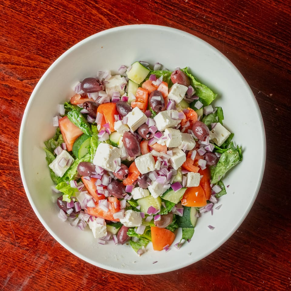 A vibrant Greek salad in a white bowl, featuring lettuce, tomatoes, cucumbers, olives, feta, and red onions.